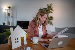 A property manager sits at a table with a laptop and a small house model, analyzing maintenance data.