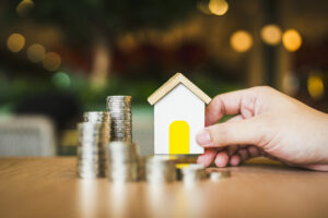 A hand holds a small house model and coins on a table, symbolizing home ownership and financial investment.