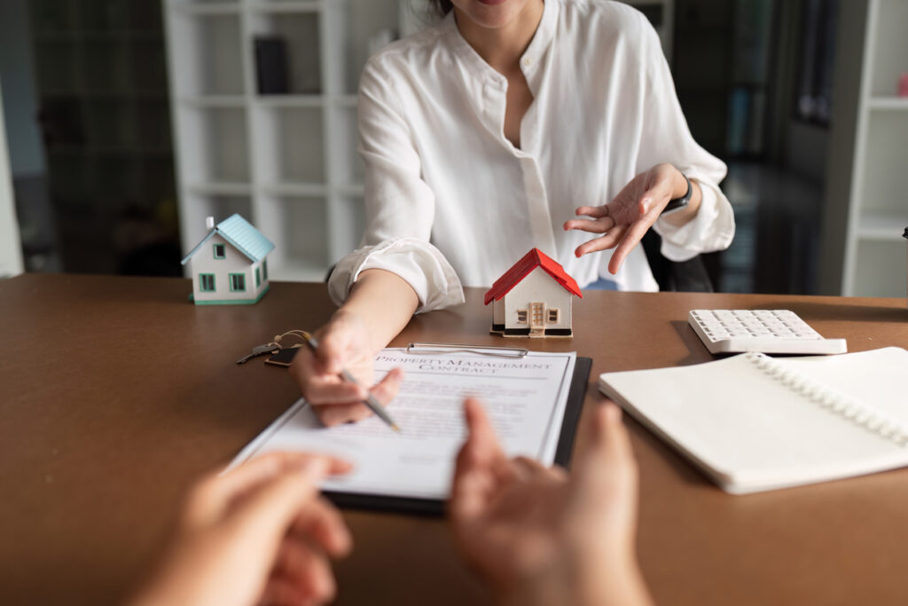 A real estate agent is signing a finders fee document in front of a house model. The woman is wearing a white shirt and the house model is red.