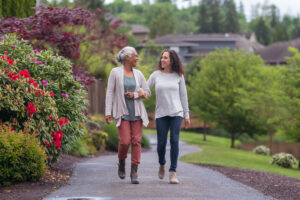 Older woman and adult daughter smiling at each other walking arm and arm through a park. 