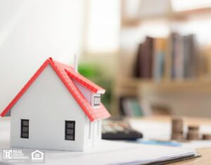A desk with a house model atop some papers, and a stack of coins in the background, highlighting property pricing.