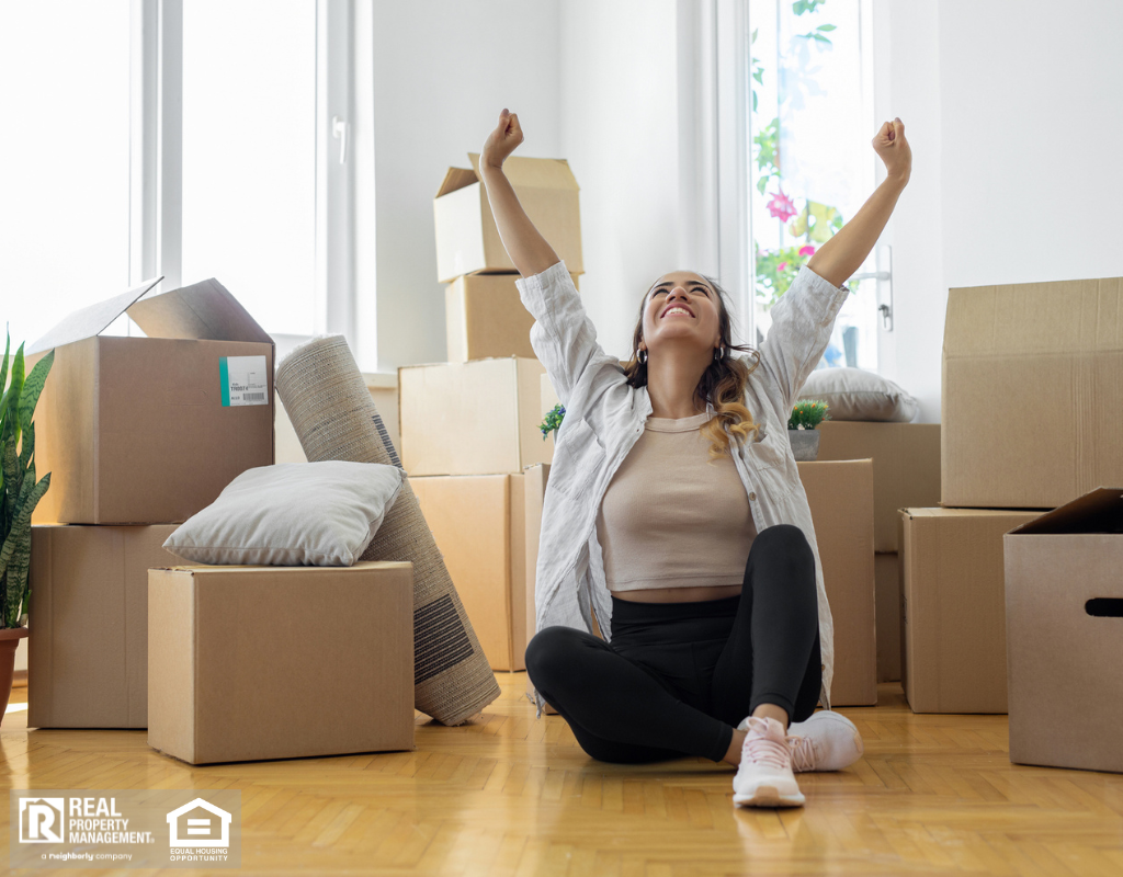 Young woman sitting on floor in new apartment with boxes and raising arms in joy.