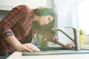 Woman in checkered shirt checking faucet while having problem with dripping faucet in kitchen.