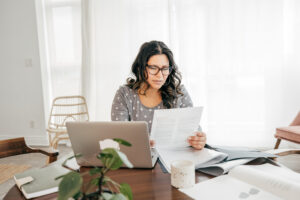 A woman wearing glasses sits at a table, working on a laptop surrounded by papers. 