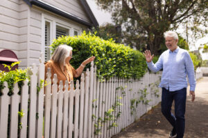 Two cheerful elderly neighbors exchange friendly greetings over a picket fence in a suburban neighborhood.