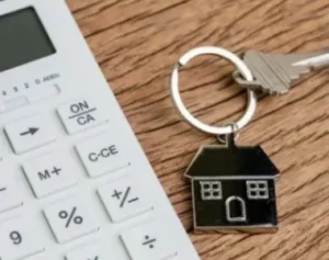 Close-up of a calculator and a house-shaped keychain with keys on a wooden surface, symbolizing real estate or home financing.
