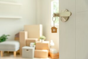 A door with a house-shaped keychain is open, showing a bright room with moving boxes and a plant in the background.