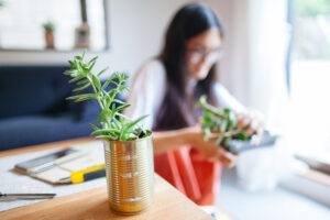 A woman seated at a table, accompanied by a plant displayed in a tin can, creating a serene indoor atmosphere.