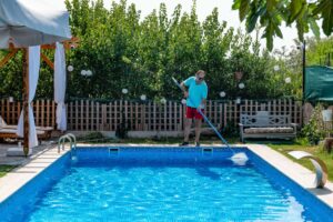 A man in flip flops cleaning a swimming pool with a net.