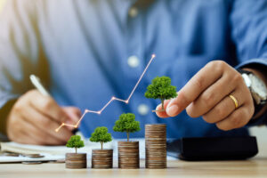 A man's hand places small trees atop stacks of coins, illustrating growth and scaling up investments.