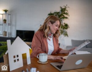 A property manager sits at a table with a laptop and a small house model, analyzing maintenance data.