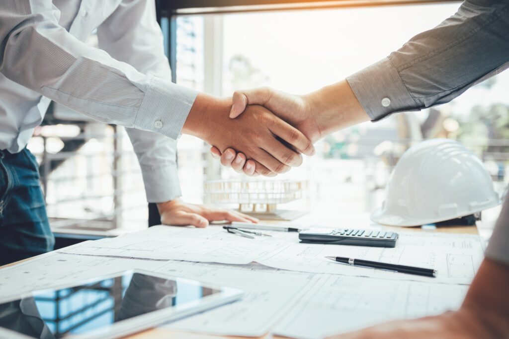 A vendor and a property manager, shake hands over a table with construction plans spread out.