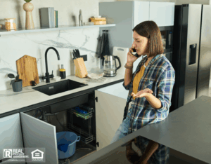 A woman in a kitchen on her cell phone, discussing an urgent maintenance issue about a leaky sink.