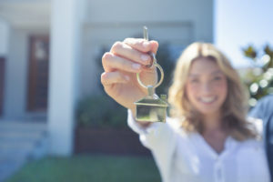 Young woman holding a house shaped key ring with a key to her new house, set in the background.