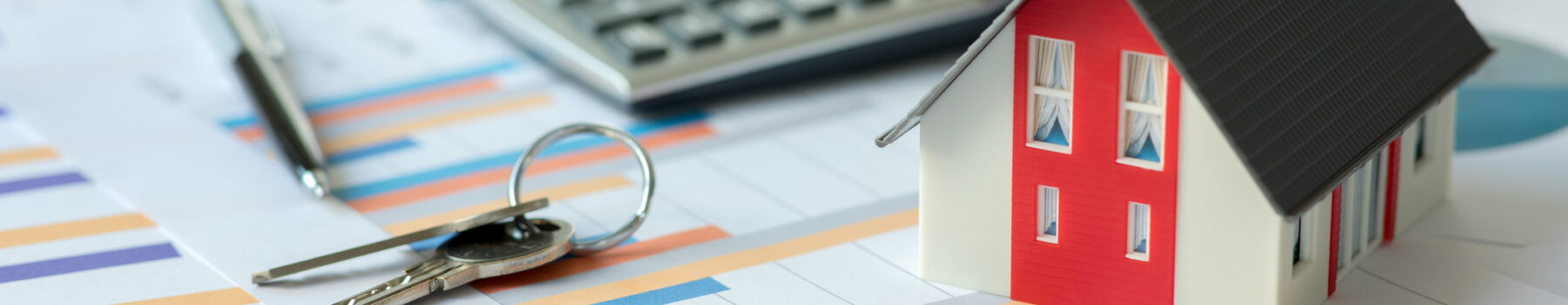Close-up of small model home on messy stack of papers with calculator, set of keys, and a pen.