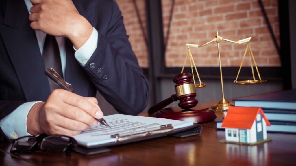 person sat at a desk that has a gavel, balance scale, and model house on it, reviewing a document