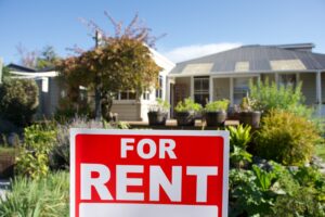 Red For Rent sign outside a house and lush garden in background.