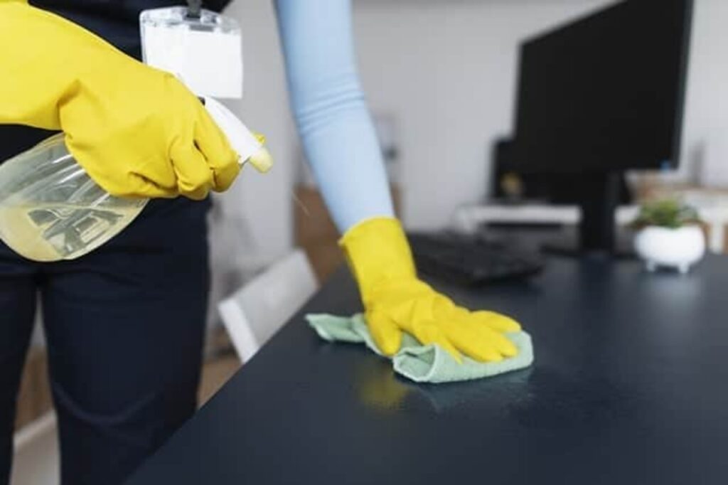 a-person-wearing-yellow-gloves-cleaning-a-desk-with-a-spray-bottle-and-cloth