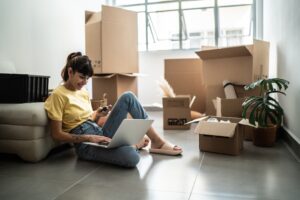 A woman on the floor with a laptop and boxes in new apartment, working on strategies to build her credit with rent payments.