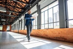 A man in a suit walks through a vacant building holding a laptop.