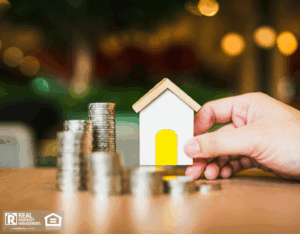 A hand holds a small house model and coins on a table, symbolizing home ownership and financial investment.