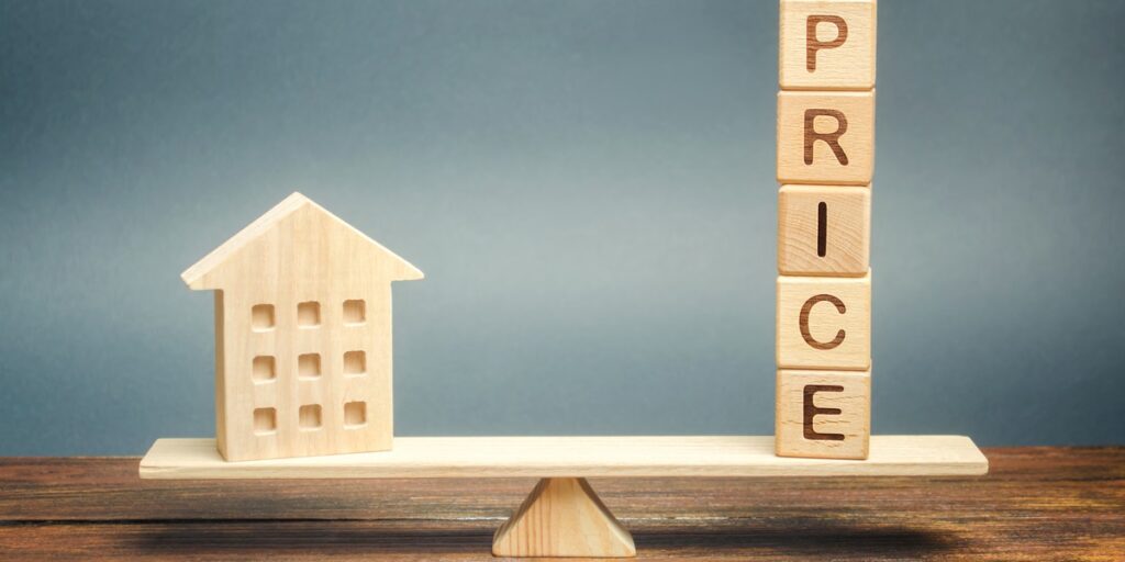 A small wooden house and wooden blocks spelling price balanced on a scale