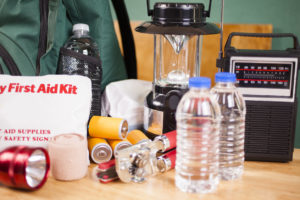 A large pile of supplies to be used in case of a natural disaster (hurricane, flood, earthquake, etc.) including: flashlight, backpack, batteries, water bottles, first aid kit, lantern, radio, can opener, mask.