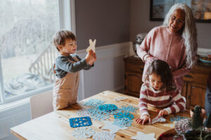 Senior woman works on a DIY winter craft with her two young grandchildren, at home in the dining room.

