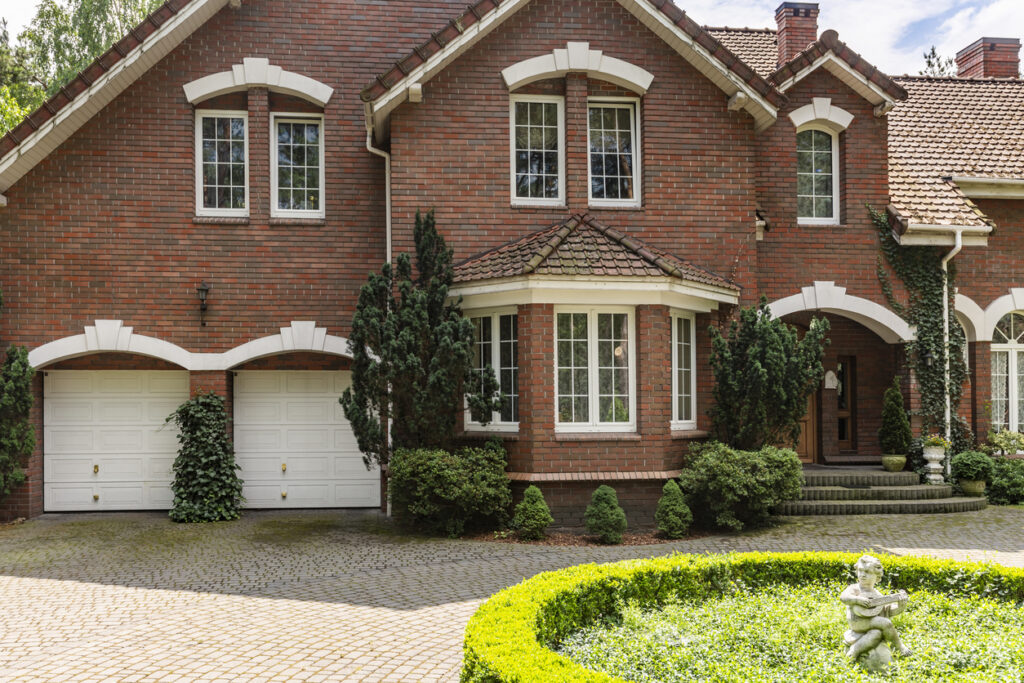 A charming older brick home with a spacious driveway and a lush garden in front.