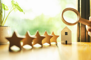 Hand of real estate businessperson holding a magnifying glass above 5 wooden stars lined up next to a wooden house model. 