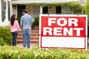 A large "For Rent" sign in front of a house with two people standing on the walkway, facing the home and holding hands.