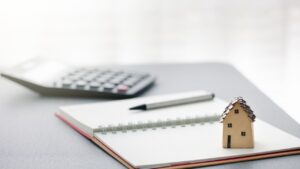 Small cardboard house beside a notebook, pen, and calculator on a desk representing property management budgeting and rental income planning.