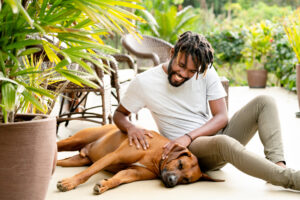A man sitting on the ground in his backyard, playing with his dog.
