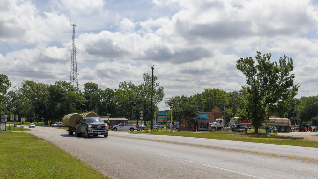 Quiet main street in Aubrey, Texas captures rural America's essence. Local businesses and residential properties line the road, while a tall communication tower dominates the skyline. Mature trees shade the street, and pickup trucks and cars parked indicate local activity