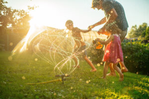 Father with children running through sprinkler at back yard in summer evening in sunset