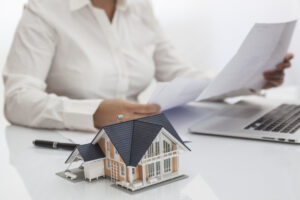 A woman working on a laptop at a desk with a house model, highlighting the advantages of real estate investment. 