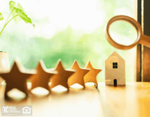 Hand of real estate businessperson holding a magnifying glass above 5 wooden stars lined up next to a wooden house model.