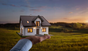 Person’s hand in foreground holding up model home in front of wide open, grassy land. 