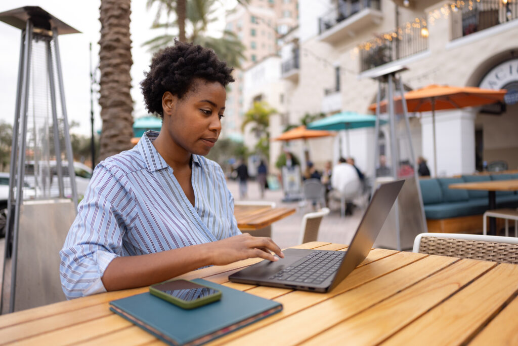 Person working on a laptop at an outdoor café with a city setting in the background.