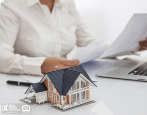 A woman working on a laptop at a desk with a house model, highlighting the advantages of real estate investment.
