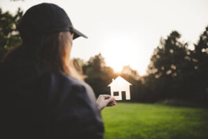 A woman holds a small paper house in her hand with sunlight shining behind it.
