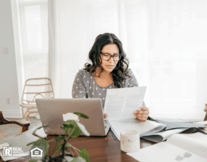 A woman wearing glasses sits at a table, working on a laptop surrounded by papers.