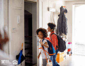 Two kids stand together in front of a door, smiling and looking at the camera on their way to school.