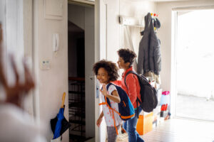 Two kids stand together in front of a door, smiling and looking at the camera on their way to school. 