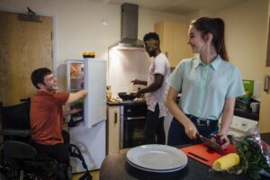 A man in a wheelchair and two other housemates collaborate in a cozy kitchen.