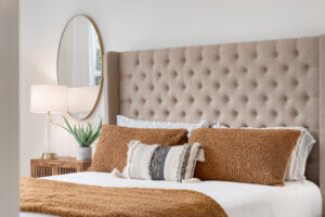 A bedroom detail shot with a cushioned headboard, a gold mirror on the wall, and a plant and lamp on the nightstand.
