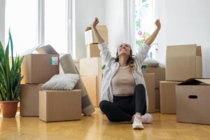 Young woman sitting on floor in new apartment with boxes and raising arms in joy.

