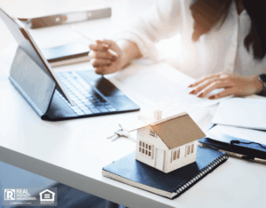 A woman uses a laptop with a house model on her desk, analyzing rental pricing strategies for rental properties.