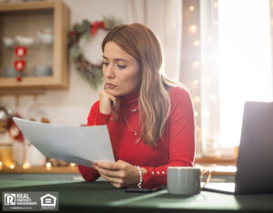 A woman wearing a red sweater sits in a holiday-decorated kitchen, intently examining a document in her hands.