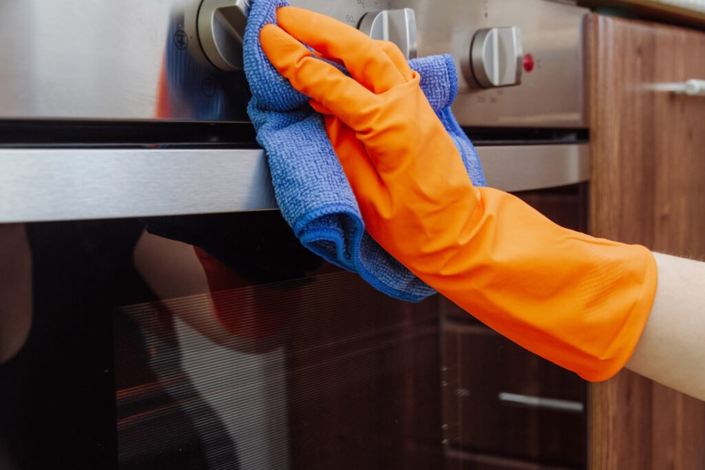 Orange-gloved hand with a microfiber cleaning cloth wiping the outside of stainless steel oven door. 